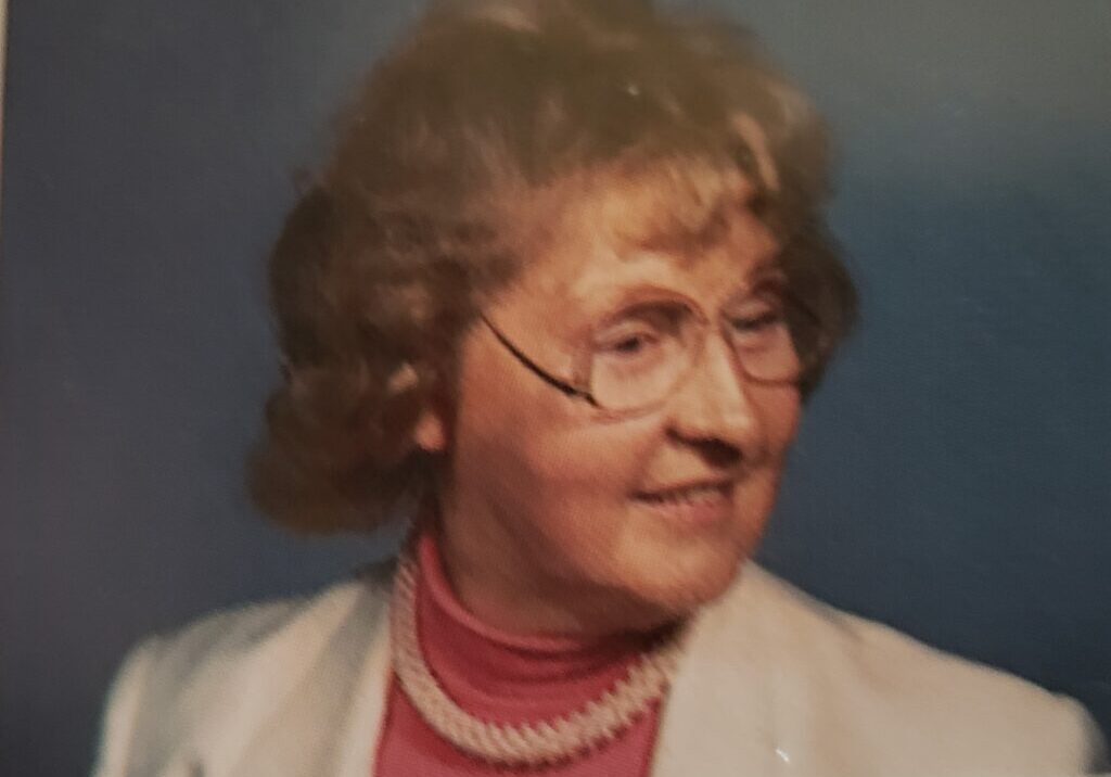 A woman with glasses and pearls in front of a blue background.